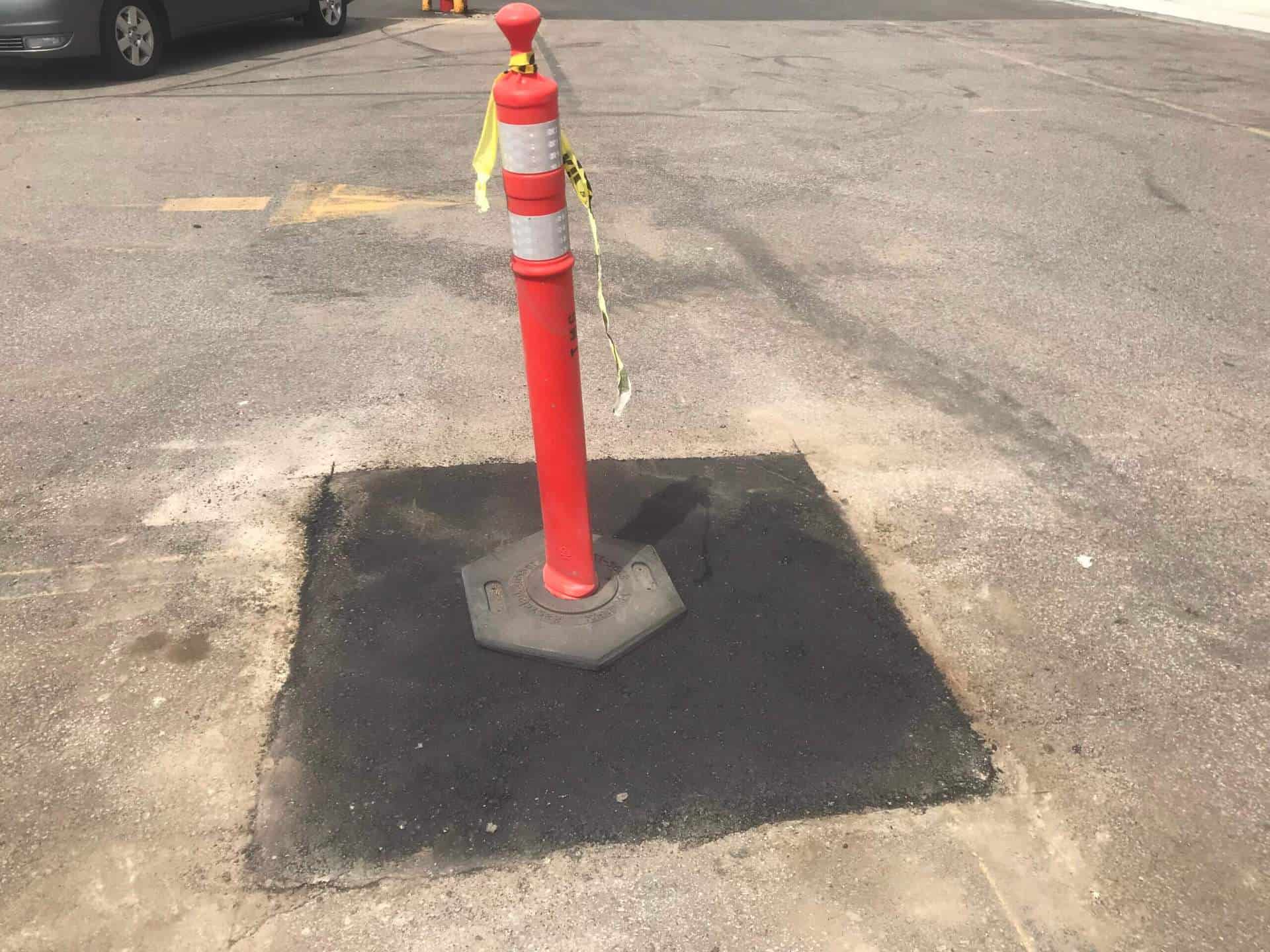 An orange construction cone sits on top of a square of new asphalt that was poured to fix a pothole.